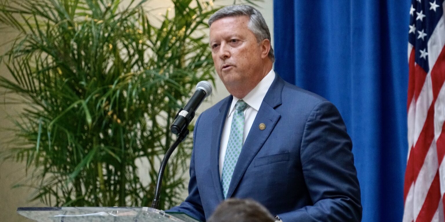 President of Jacksonville University with a green tie and blue suit in front of a podium speaking. There is an American flag to the right, and to the left is an artificial palm. 