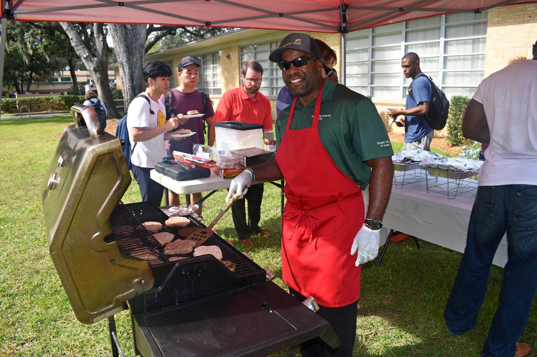 African American male in red apron cooks food for veteran students outside 