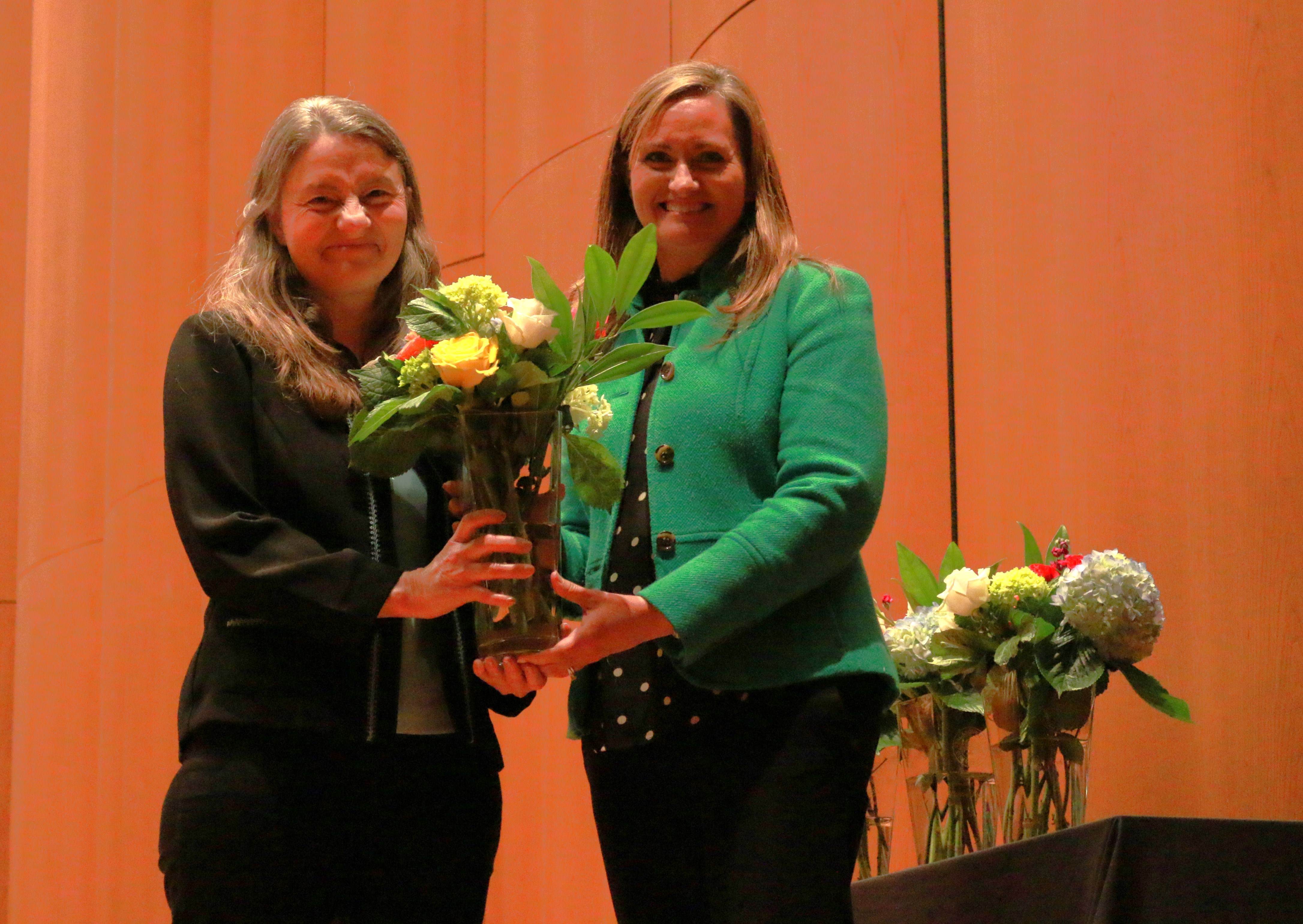 Two women standing beside eachother, one holding a vase of flowers