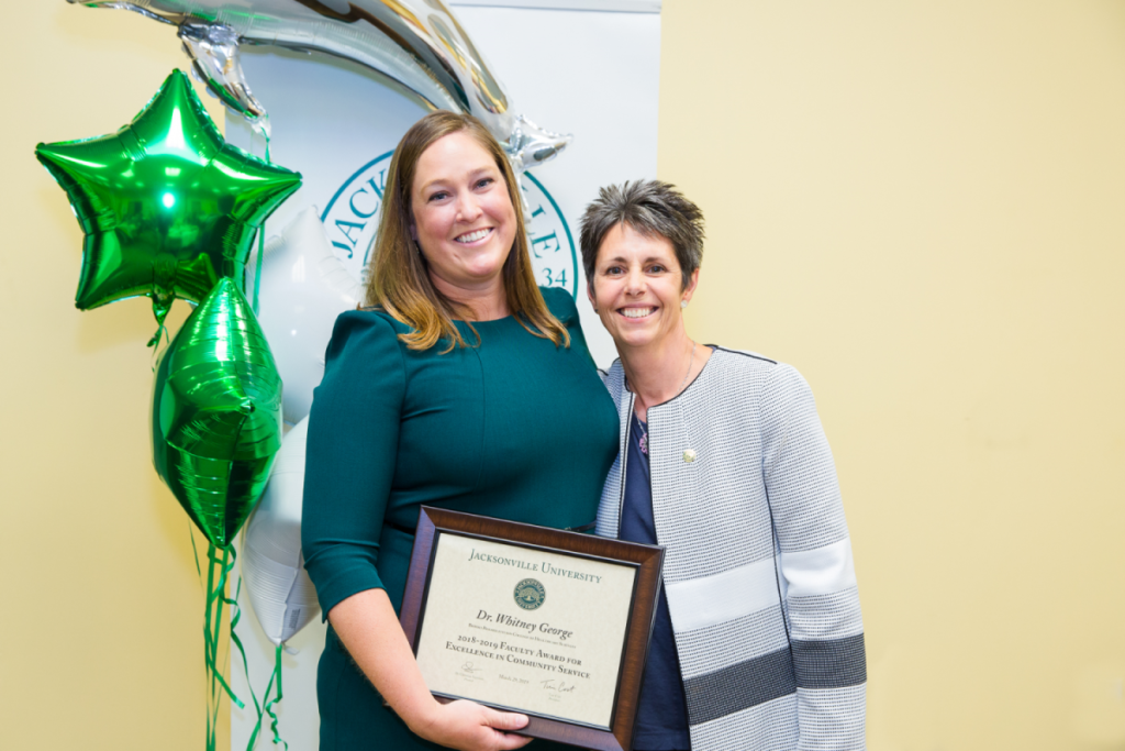 Whitney George and Dr. Christine Sapienza pose and smile for a photo while Whitney holds up her award plaque in front of a JU banner and green star balloons