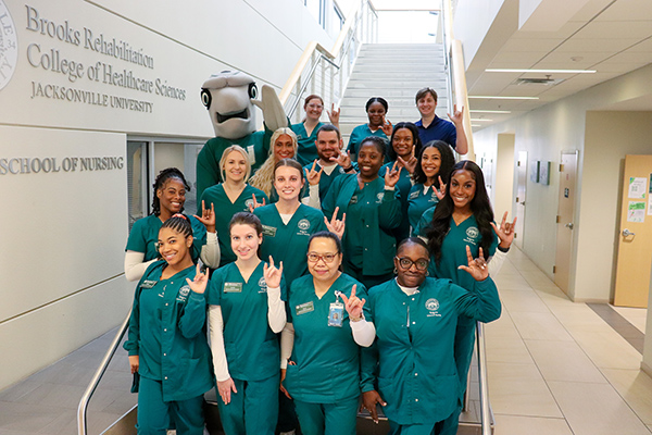 Cohort of ABSN students pose with Dunk'n the Dolphin on the steps of the Keigwin School of Nursing staircase.