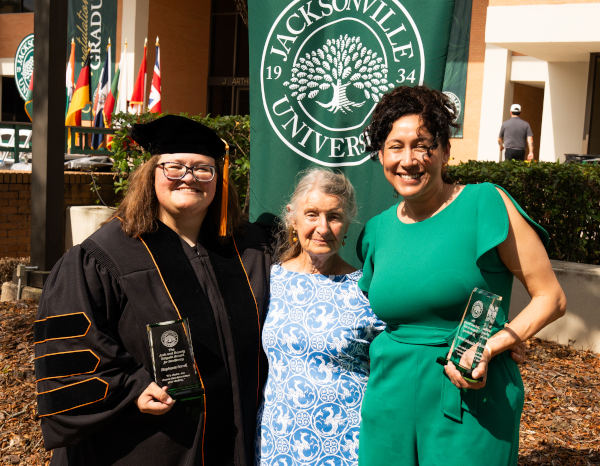 Three women smiling outdoors in front of a Jacksonville University banner, including a graduate in cap and gown holding an award and another woman holding a glass plaque.