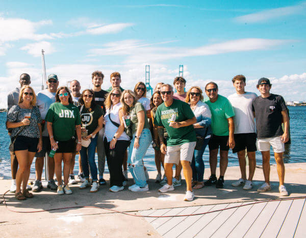 Group of about twenty adults and young adults standing together on a waterfront dock on a sunny day, smiling toward the camera. Several people wear green shirts with college-style logos, and some hold drinks. Blue water and tall port cranes appear in the background under a bright sky with scattered clouds.