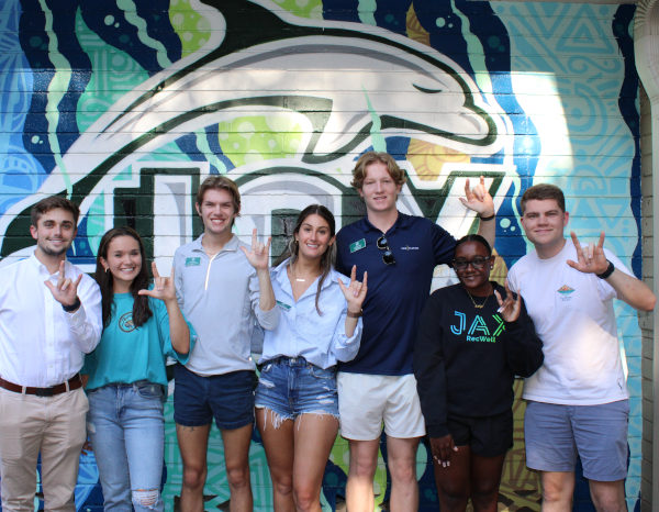 A group of students posing in front of a dolphin wall mural with their hands in the Phins Up sign.