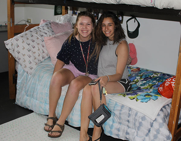 Two female students sit on a bottom bunk together and smile for a picture during move-in.